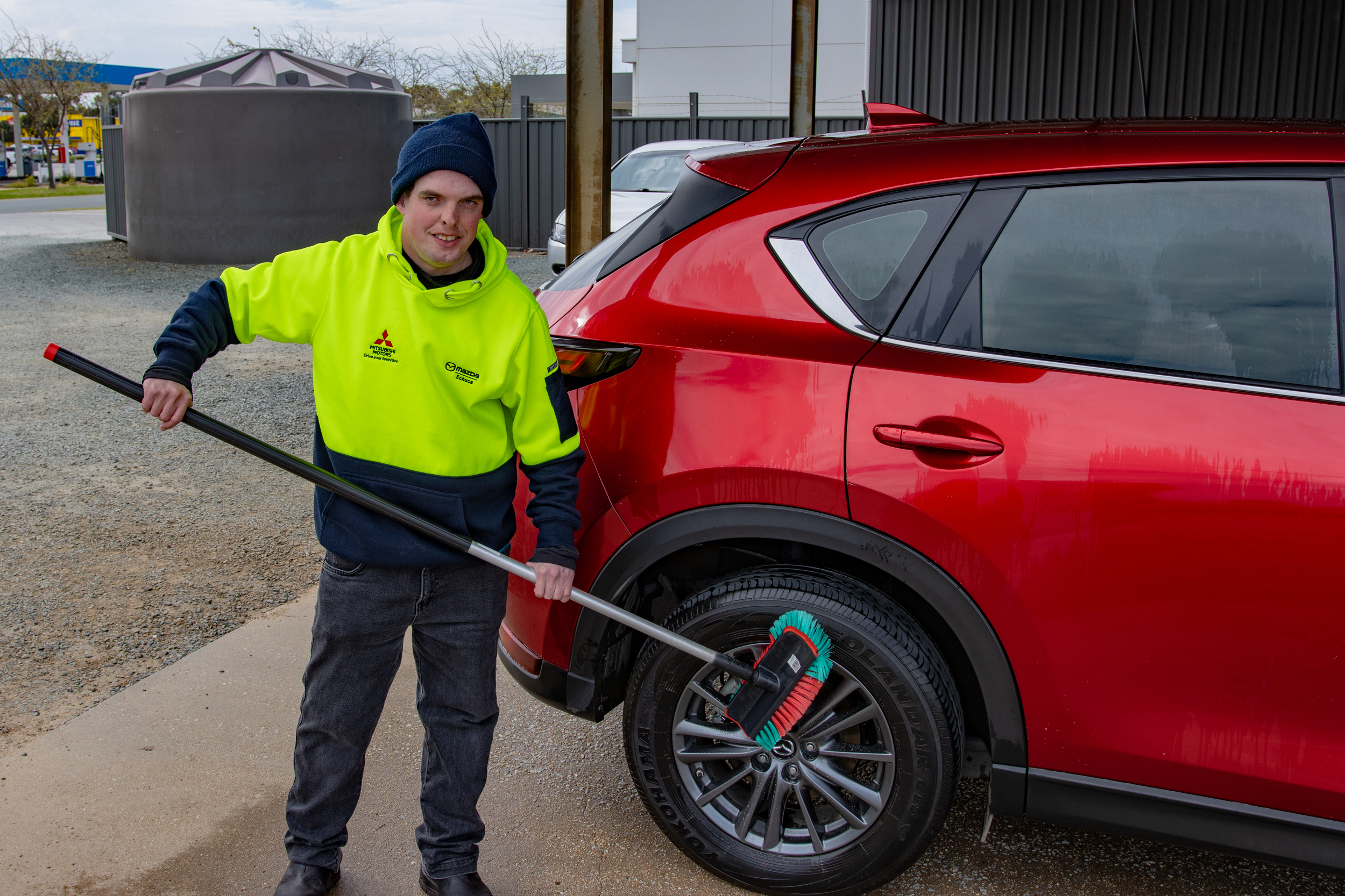 Young man in beanie and yellow jumper smiling as he holds a car cleaning brush that he is using to clean a red SUV.