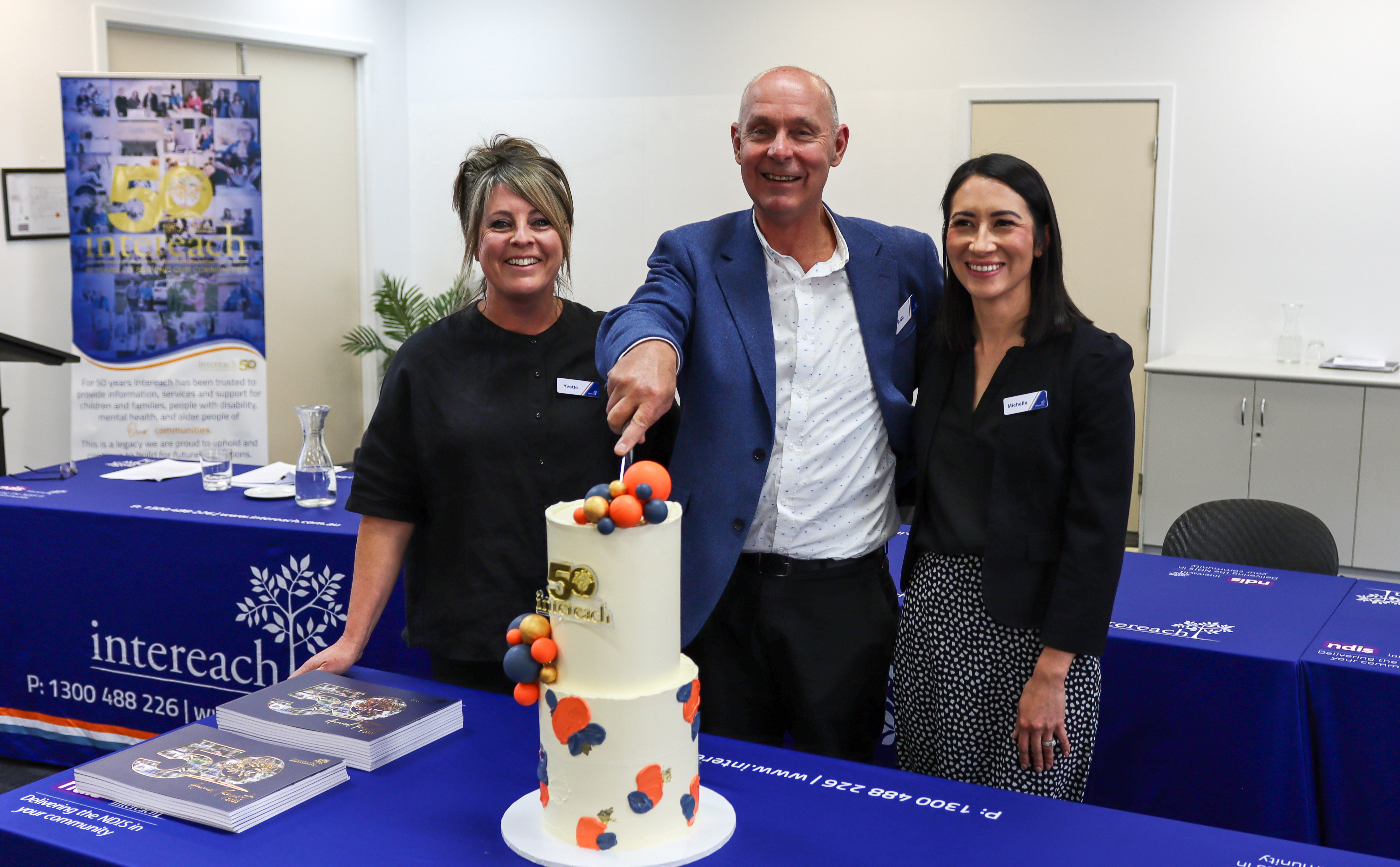 Two women dressed in black and a man in a suit standing in a board room behind a table smiling while cutting a two-tiered cake to celebrate Intereach's 50th anniversary