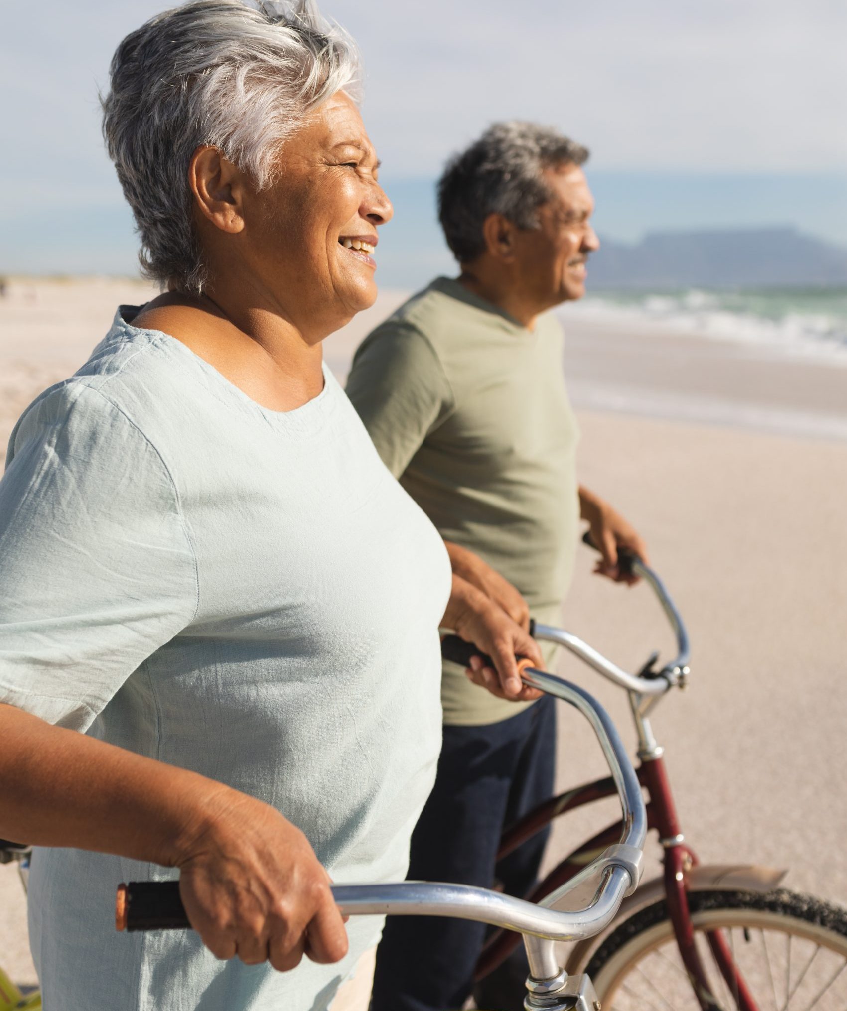Side view of smiling senior multiracial couple looking away with bikes on shore at sunny beach. active lifestyle and transportation.